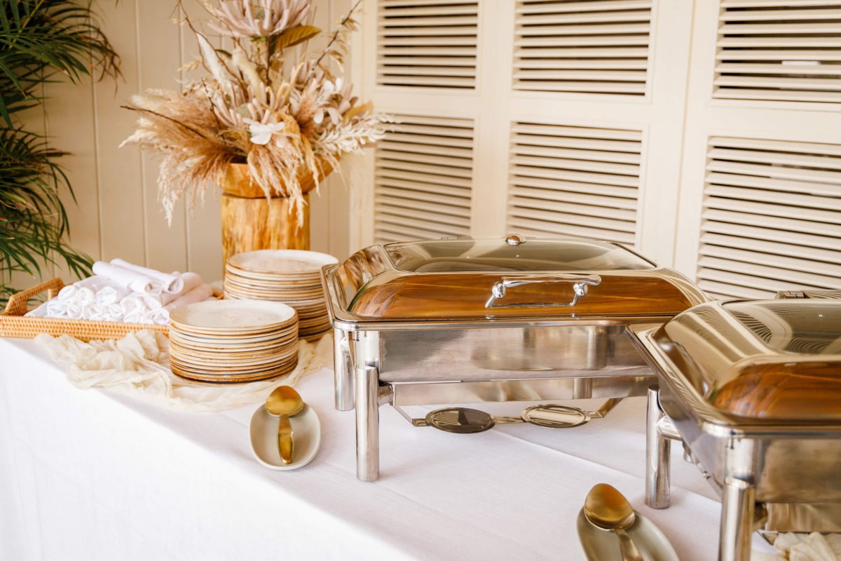 A table with metal food serving containers, stacks of plates and cutlery