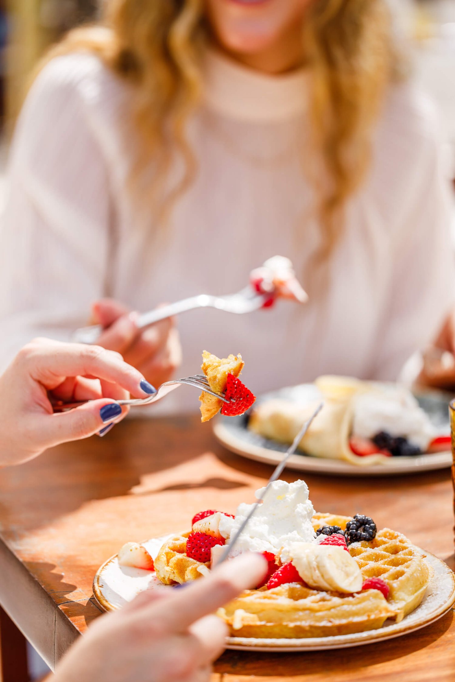 Two women sitting at a dining table, eating waffles and pancakes topped with cream, strawberries and banana