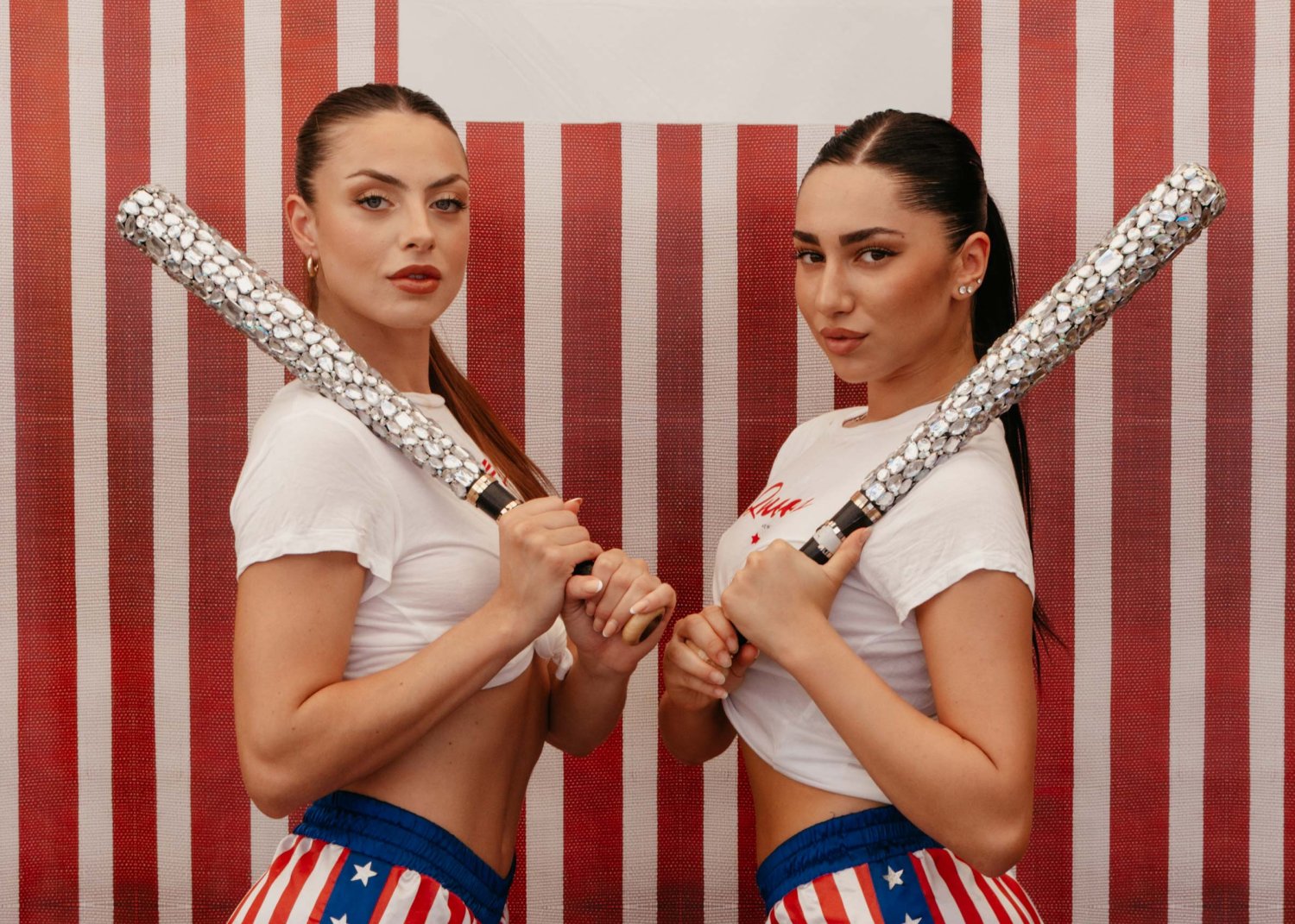 Two women posing in American flag clothing, holding sparkly baseball bats