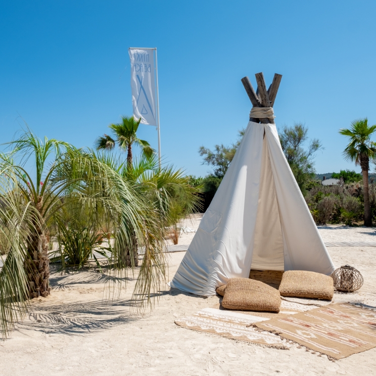 A teepee with cushions and blankets laid in front. A flag post of the Nikki Beach logo is beside the teepee as well as small palm trees