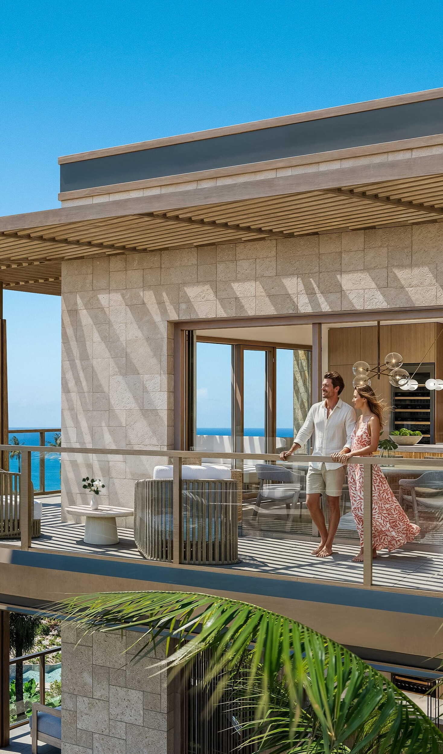 Exterior view of a modern luxury condominium featuring a spacious curved balcony with glass railings. A couple stands on the terrace enjoying a panoramic view of the bright blue ocean, surrounded by tropical palm trees and landscaped gardens under a clear blue sky