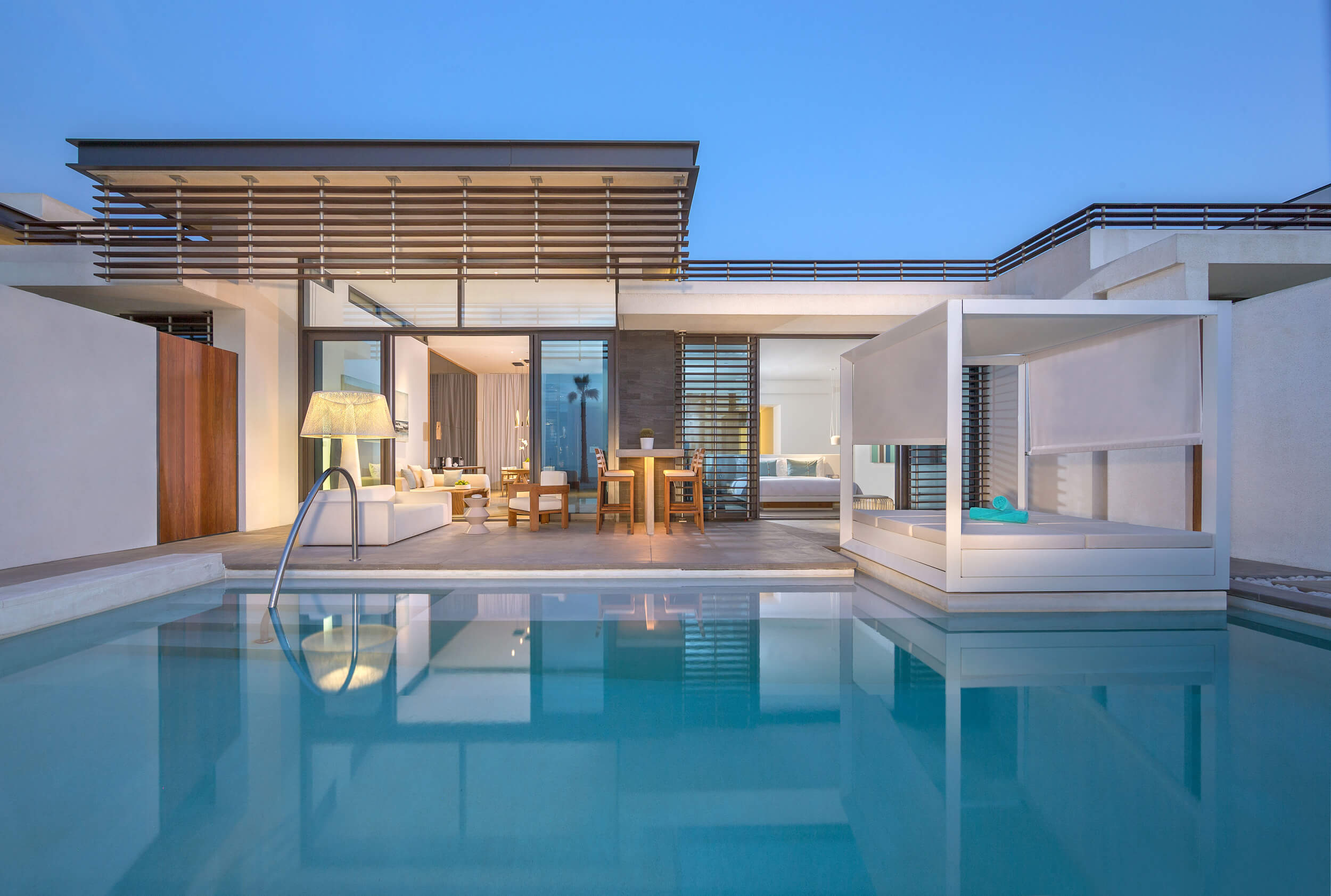 A view across the pool of the Beach villa, showing the sun bed, outside seating areas. Sliding doors are open, showing the living room areas inside