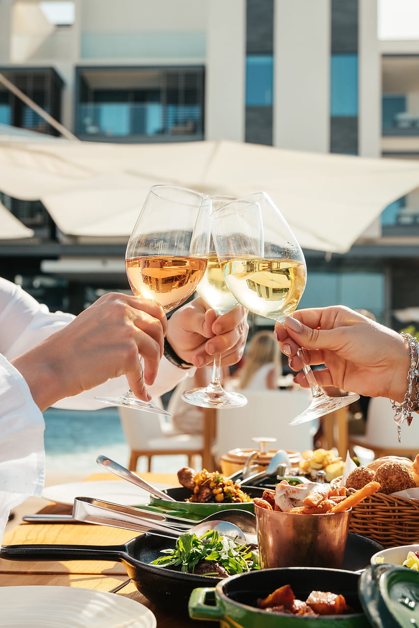 A close-up of three friends toasting with glasses of white and rosé wine over a wooden table filled with an appetizing spread of dishes served in cast-iron skillets and a copper cup. The blurred background features white sun umbrellas and bright sunshine, perfectly capturing the chic beachside lunch experience at Café Nikki