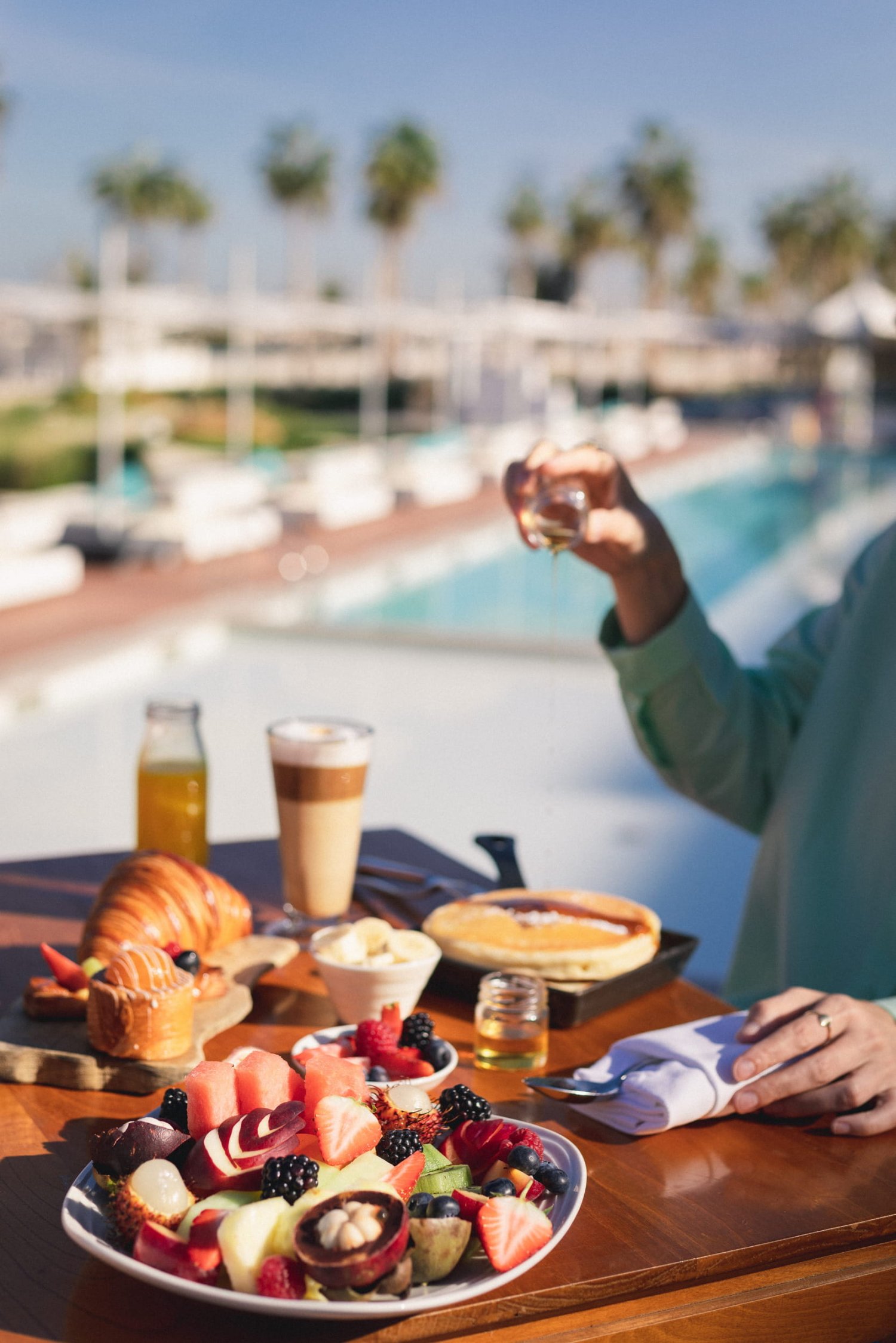 A close-up of a vibrant breakfast spread on a wooden table, featuring a large plate of freshly cut fruit, pastries, an iced coffee, and a person's hand pouring syrup over a pancake. The brightly lit, blurred background reveals a pristine swimming pool lined with white sunbeds and palm trees, perfectly illustrating the transition from a Café Nikki morning to a relaxing day by the water