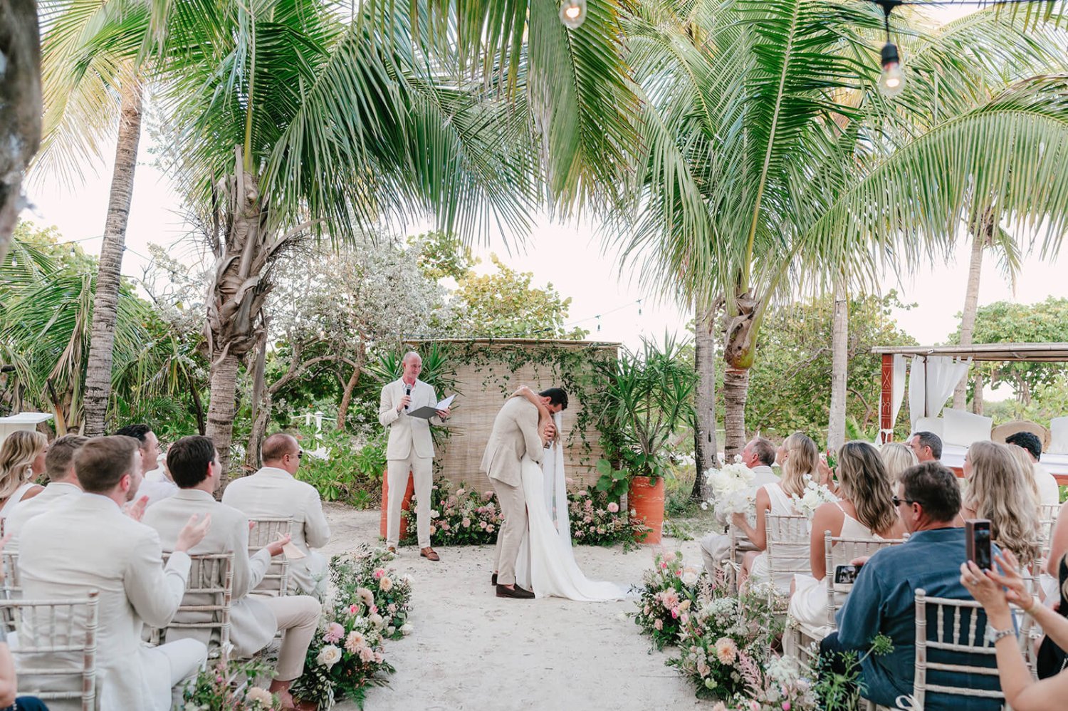 A couple hugging after getting married on a beach, surrounded by family with palm trees in the background