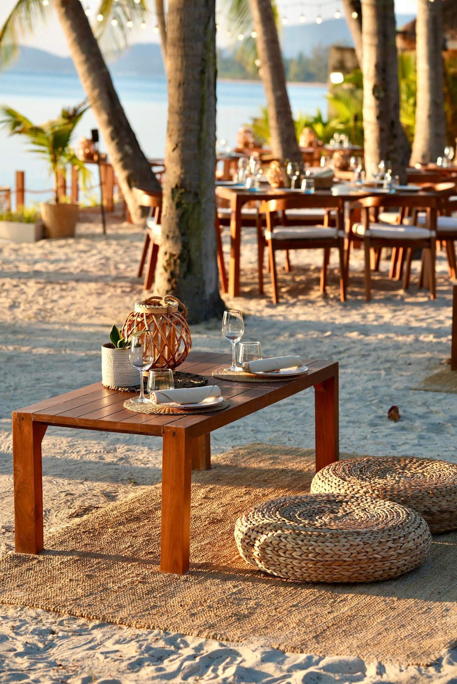 A sandy beach with a small table and stools that sit close to the floor, looking out to the sea. Dining tables and chairs in the background