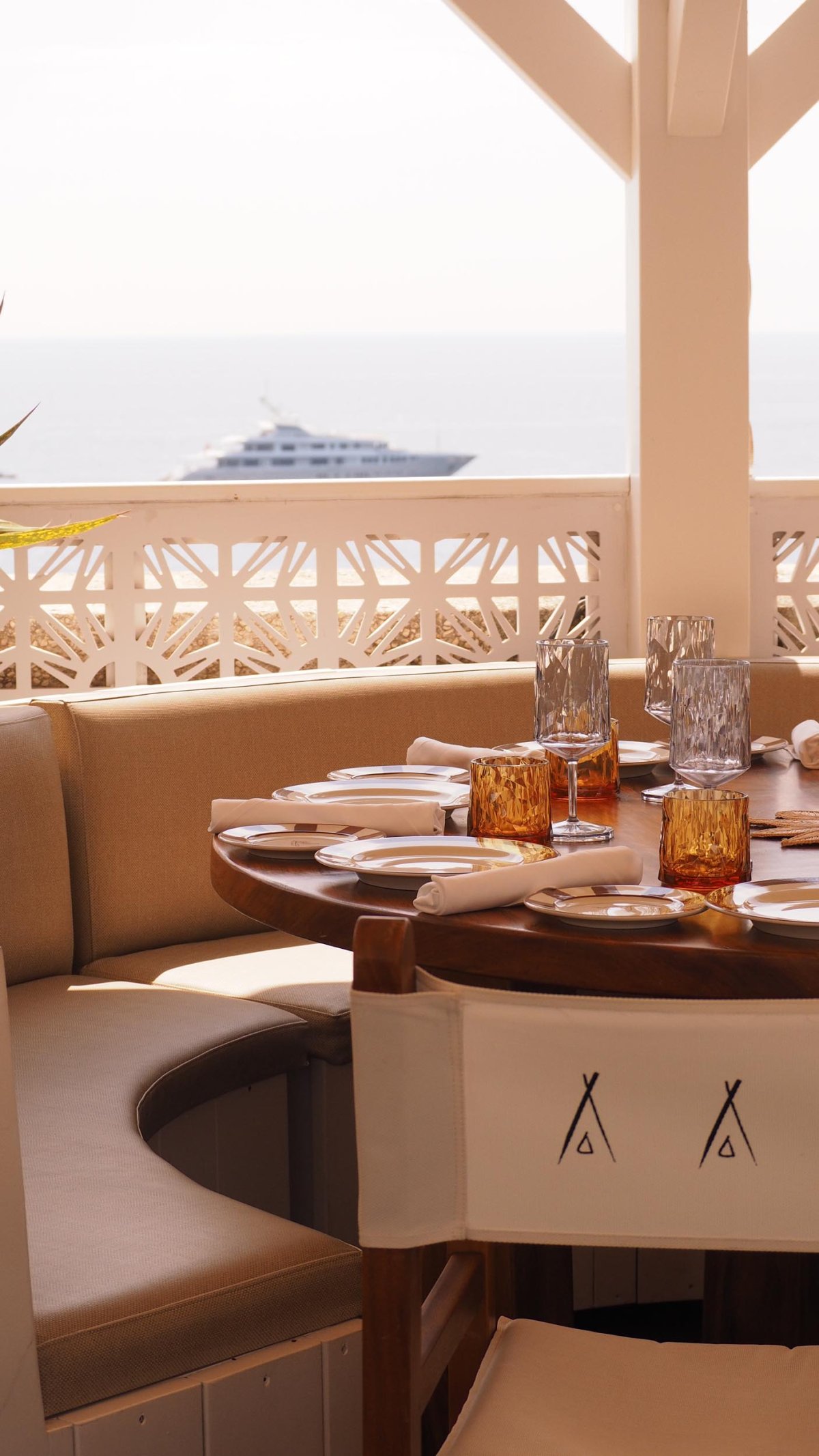A table set with plates, glasses and cutlery. Behind is a sea view with a passing boat
