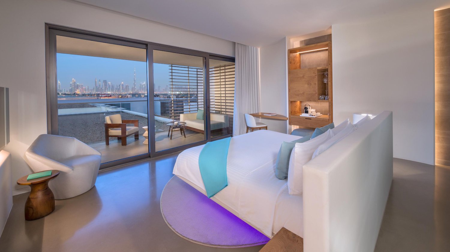 King sized bed with a small counter of glasses, water and coffee. The bed looks out the the skyline view balcony