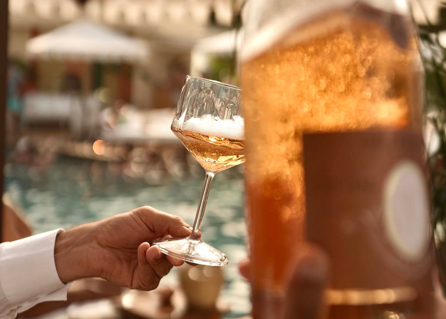 A person holding a glass of sparkling wine with a bottle held at the front of the frame