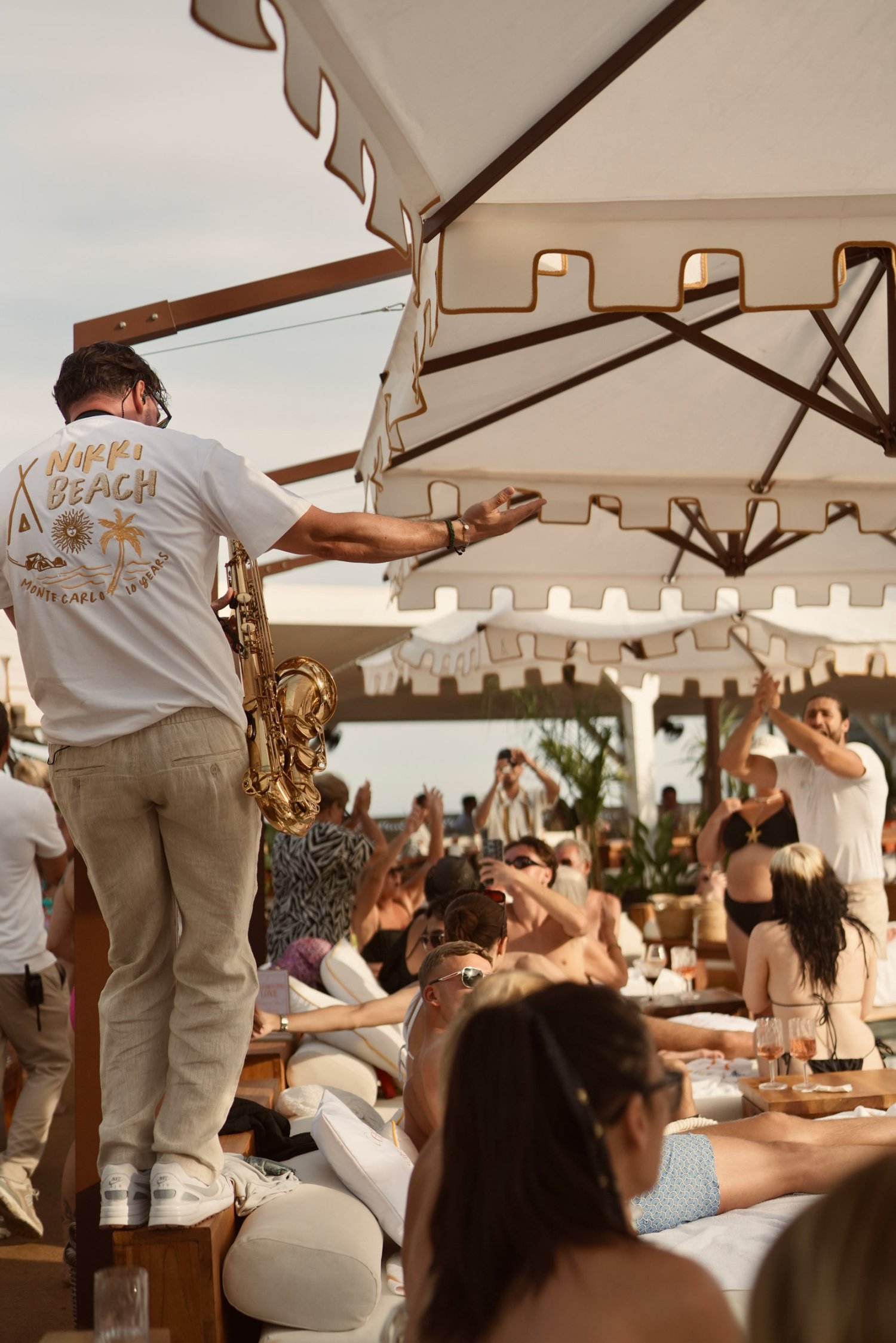 A man standing on a table, playing the saxophone to a crowd of people at Nikki Beach