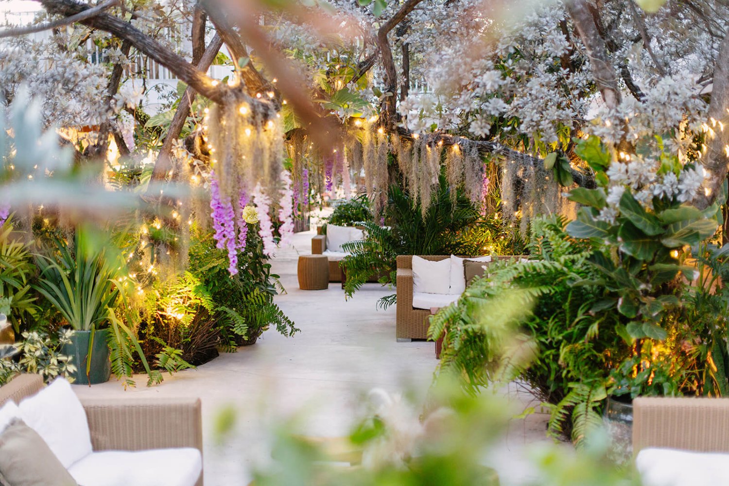 The garden - Nikki Beach Miami. A bright scene with archways of flowers and greenery surrounding lounge chairs and coffee tables. Fairy lights are intertwined with the branches of over hanging trees