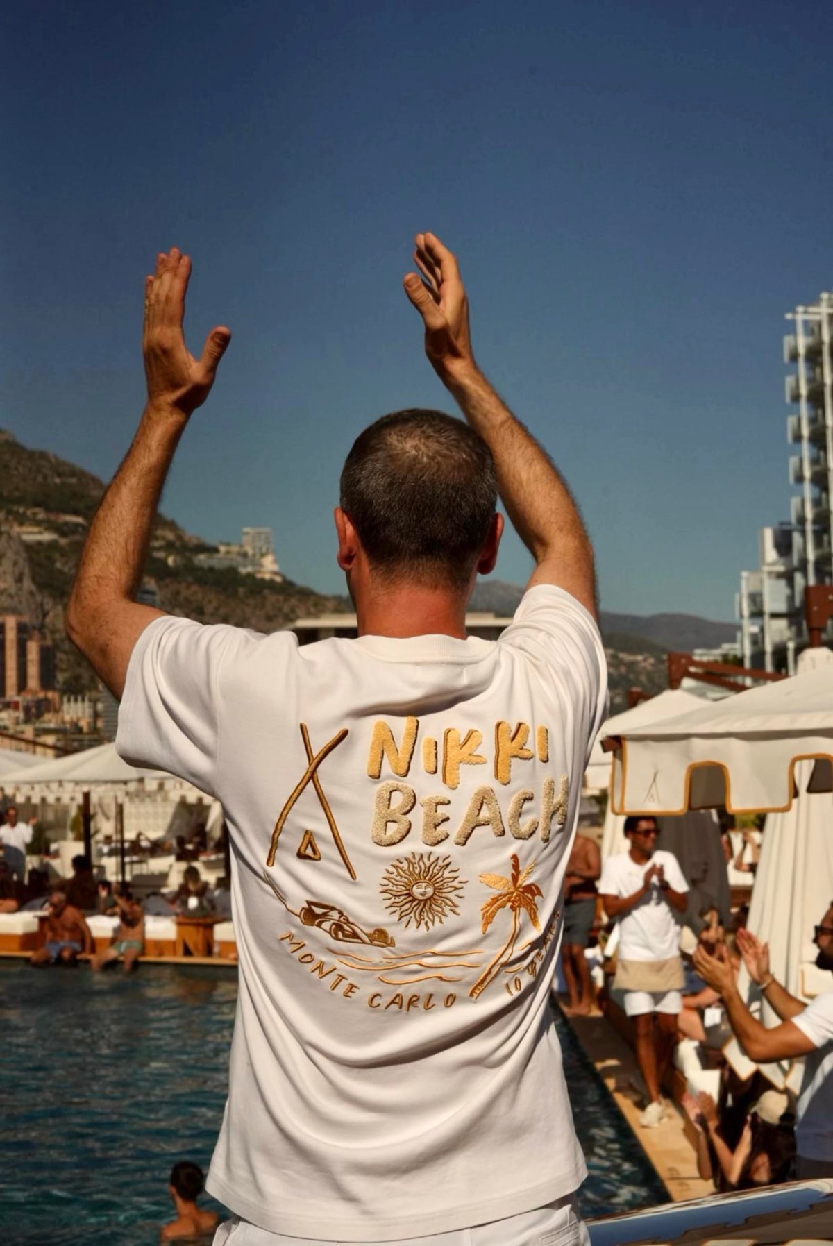 A man wearing a Nikki Beach t-shirt, clapping his hands above his head