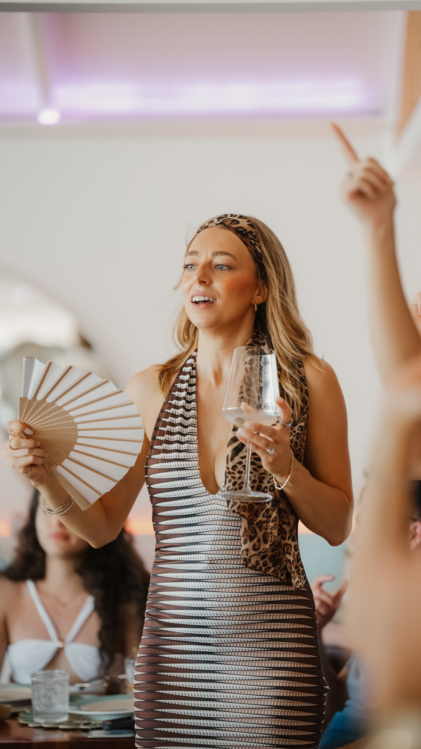 A lady holding a glass of wine and holding a fan in the other waving it to cool herself down