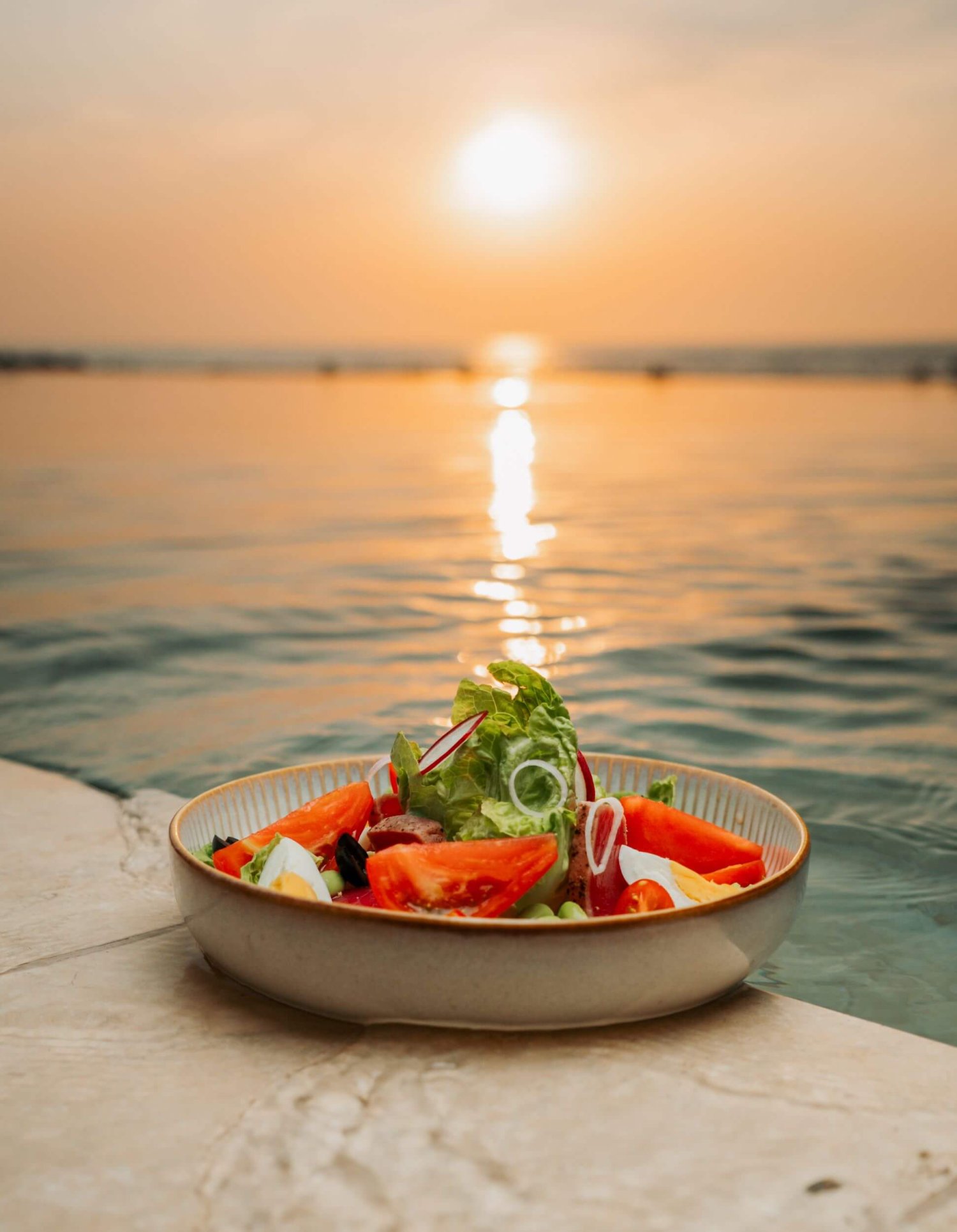 A colorful bowl of salad featuring fresh tomatoes and lettuce sits by a pool, bathed in the warm glow of sunset at Nikki Beach Baku