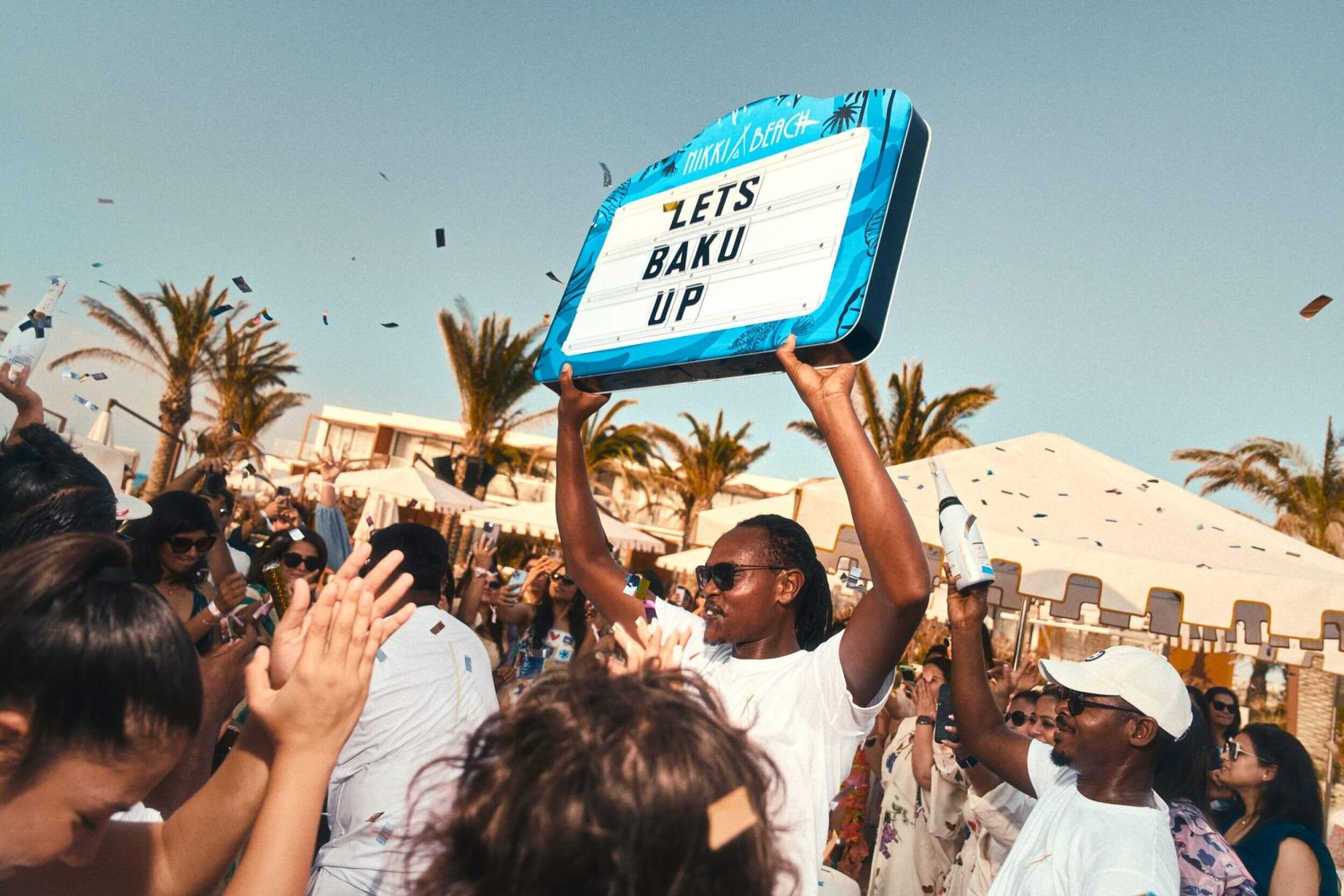 A vibrant crowd celebrates outdoors with confetti falling through the sunny sky. At the center of the energetic party, a man in sunglasses enthusiastically holds a large, illuminated blue sign reading 