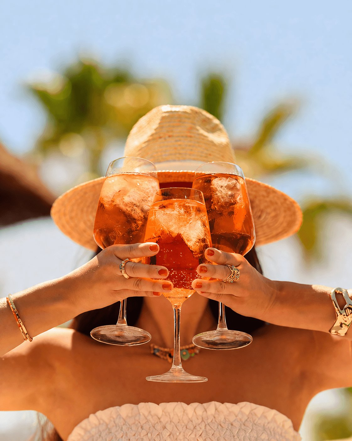 A person wearing a wide-brimmed straw hat playfully holds three large, vibrant orange Aperol Spritz cocktails in front of their face. The bright, sunlit background with blurred palm trees perfectly captures the refreshing 