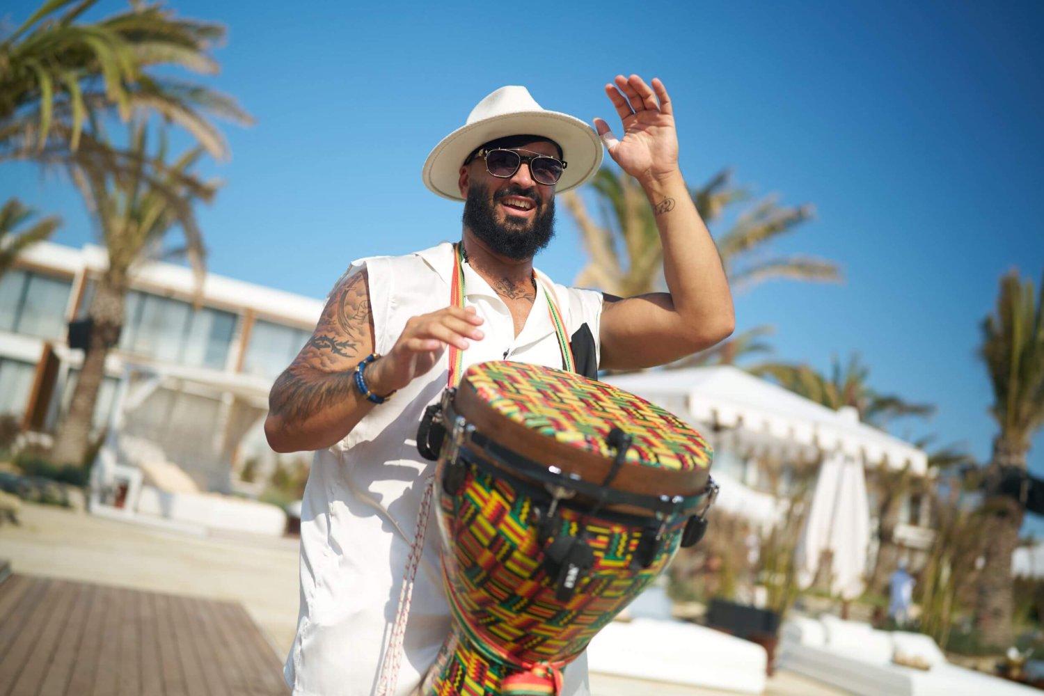 A smiling live drummer wearing a white hat and sunglasses energetically plays a vibrantly patterned drum outdoors. With one hand raised mid-beat against a bright blue sky and a backdrop of palm trees and white beach cabanas, this scene perfectly captures the upbeat, seaside rhythm and captivating live performances of the weekly Friday Sun Sessions