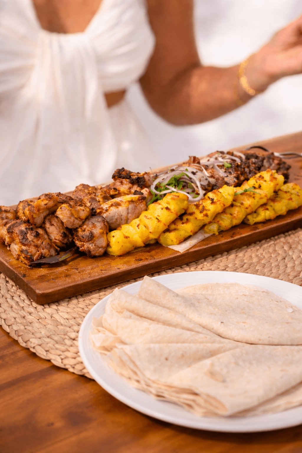 A close-up view of a vibrant sharing board loaded with grilled meat skewers and spiced potatoes, garnished with fresh herbs and thinly sliced red onions. Accompanied by a plate of folded flatbread with a guest in white resort wear blurred in the background, this mouth-watering spread perfectly showcases the bold local Azerbaijani flavors and social dining experience of the weekly Taste of Baku Thursday event