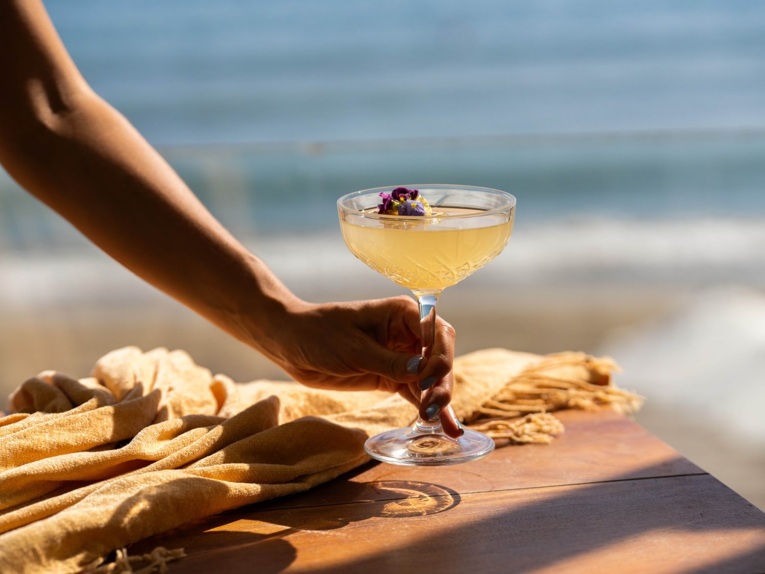 A hand elegantly holds a vibrant cocktail atop a polished wooden table, exuding a sense of luxury and relaxation