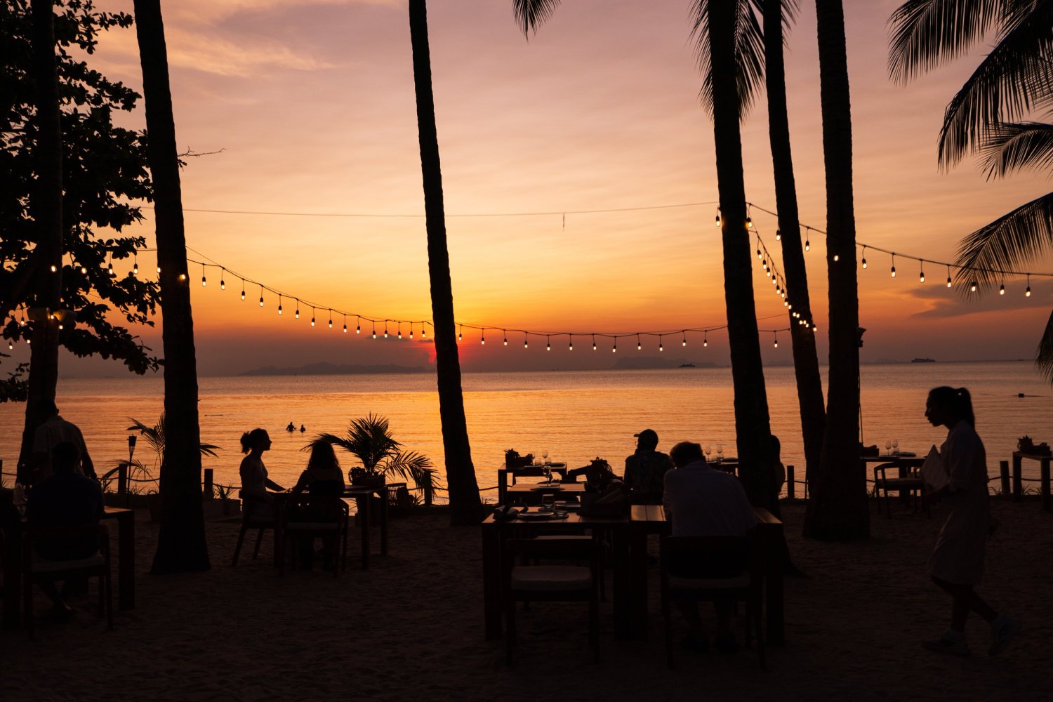 A group of elegantly dressed people enjoying a leisurely meal at tables set on a picturesque beach
