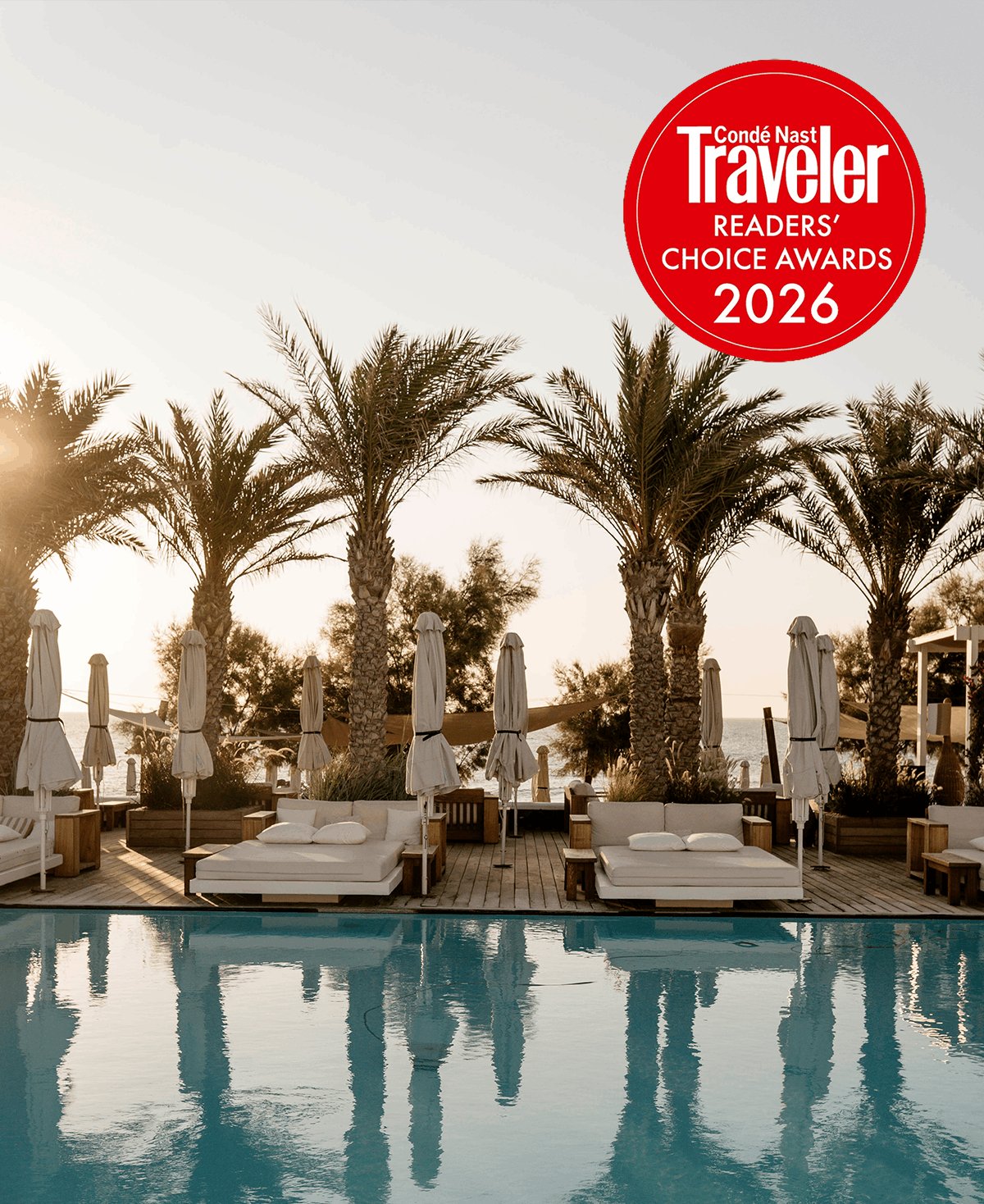 A tranquil golden hour view over a swimming pool lined with a wooden deck, plush white sunbeds, closed umbrellas, and lush palm trees reflecting in the water. A red Condé Nast Traveler Readers' Choice Awards 2026 badge sits in the top corner to thank guests for their nominations