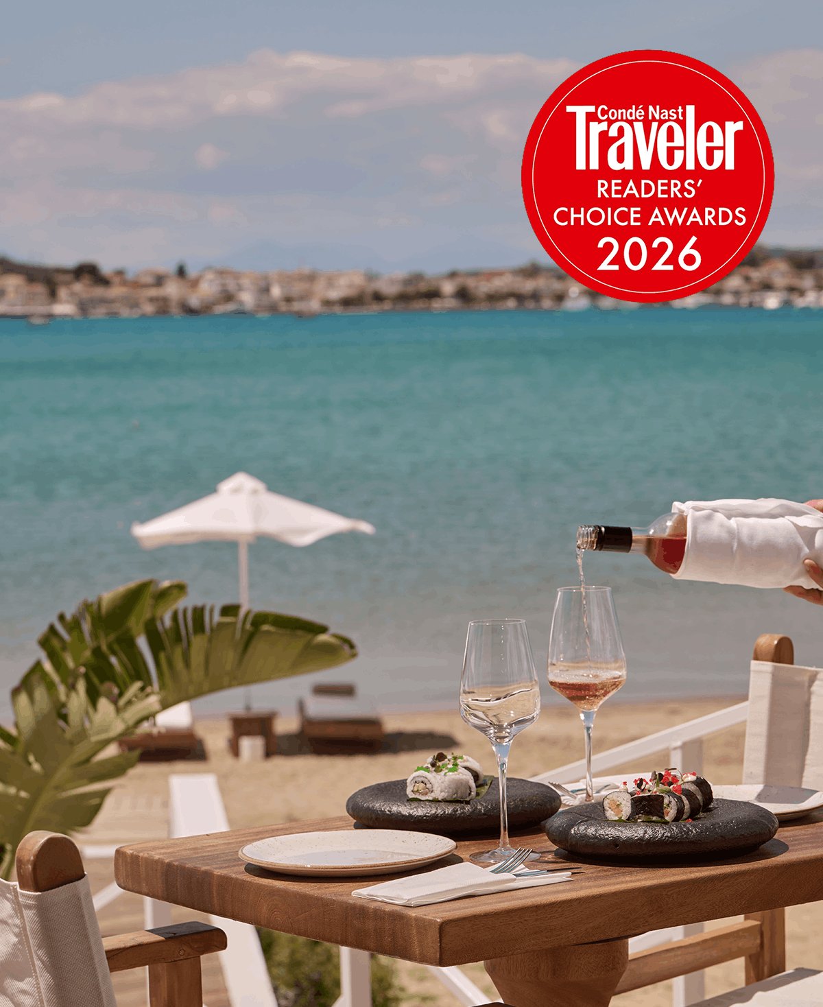 A close-up of a seaside dining table where a hand pours rosé wine into a glass, accompanied by beautifully plated sushi on black stones. The bright blue sea sits in the background, and a red Condé Nast Traveler Readers' Choice Awards 2026 badge is displayed in the top corner to encourage guest votes