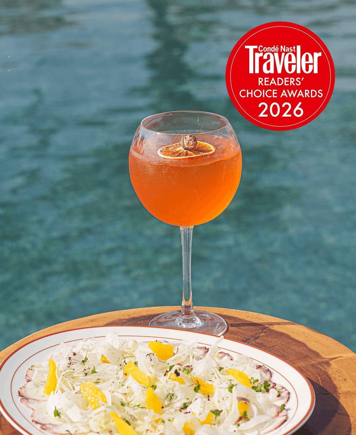 A vibrant orange spritz cocktail garnished with dried citrus rests on a wooden table next to a plate of fresh fennel and citrus salad. Sparkling blue water fills the background, with a red Condé Nast Traveler Readers' Choice Awards 2026 badge in the top right