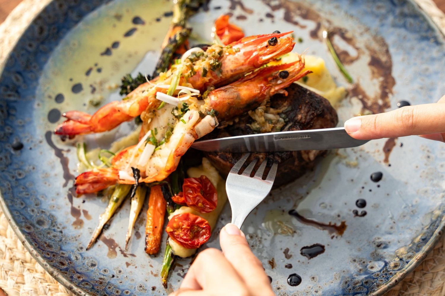 A person elegantly holds a knife and fork above a beautifully plated BBQ dinner, ready to savor the flavors of Thai cuisine