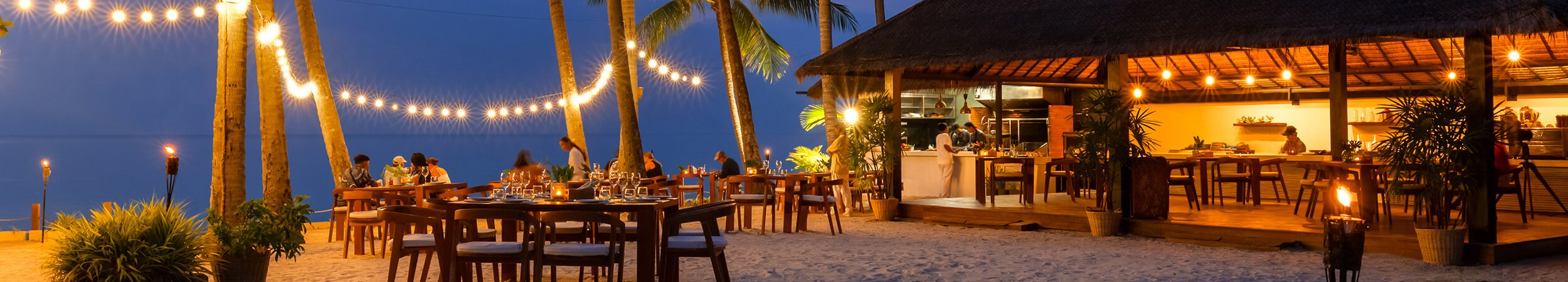 A picturesque beach at dusk, showcasing a delightful restaurant and graceful palm trees framed by a vibrant sunset sky