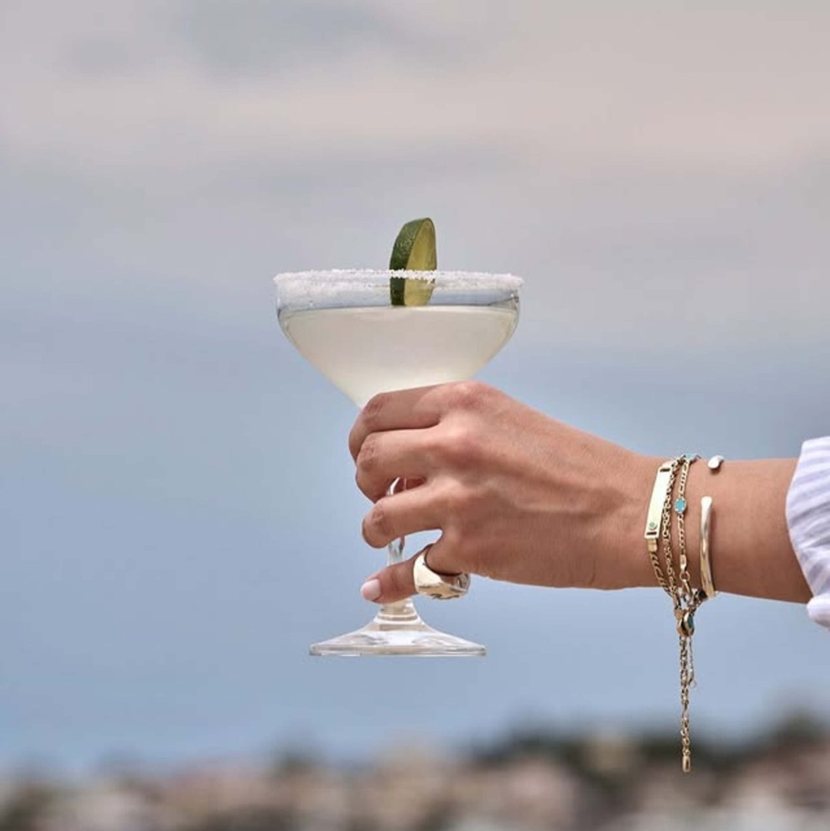 A chilled cocktail lifted against the horizon — sunlight glinting off the glass, sea breeze swirling through golden bracelets
