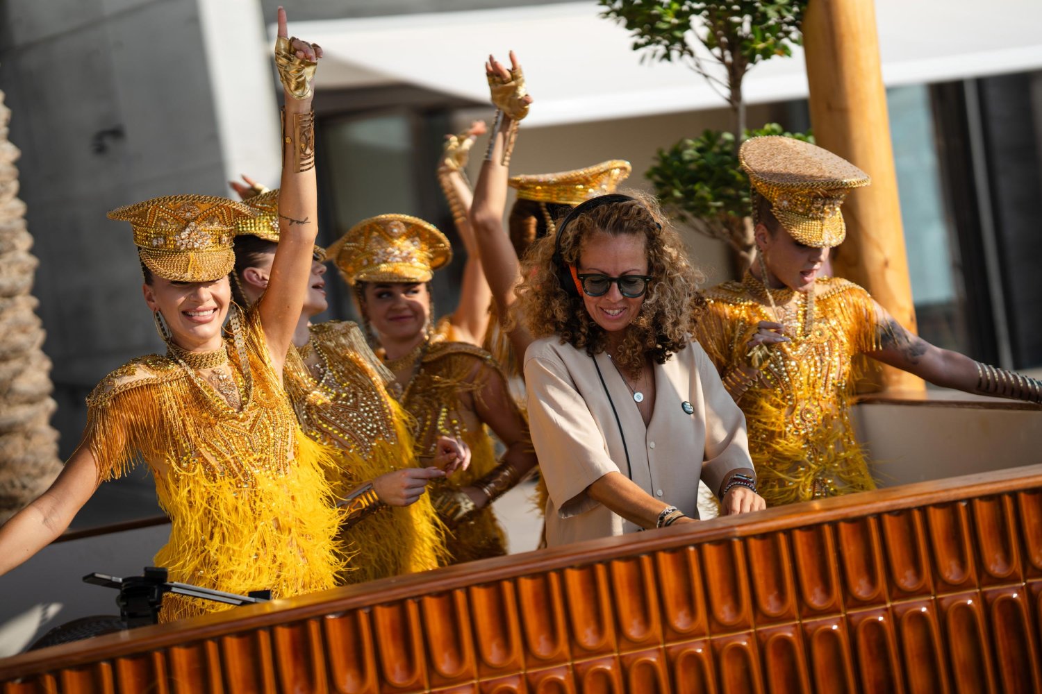 A qomen mixing on DJ decks with people dancing behind in vibrant costumes