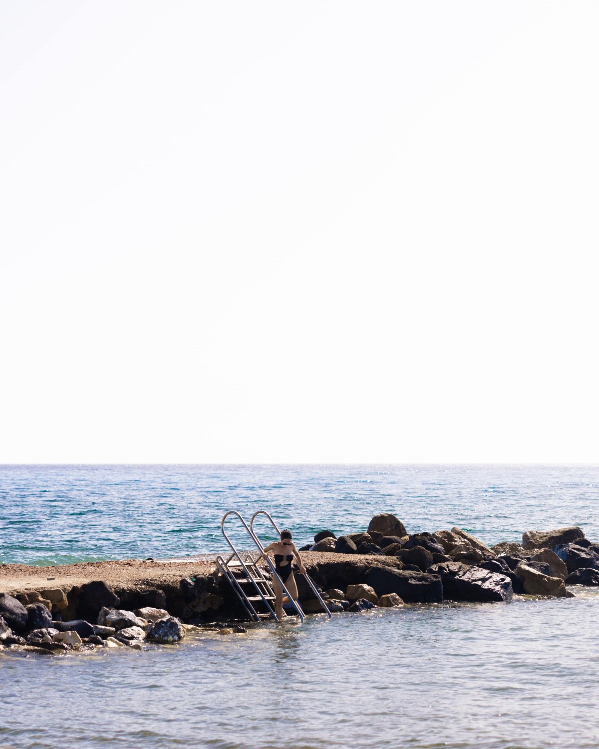 A quiet descent into the clear blue, where the rocks meet the rippling Aegean. A moment of solitude framed by endless horizon