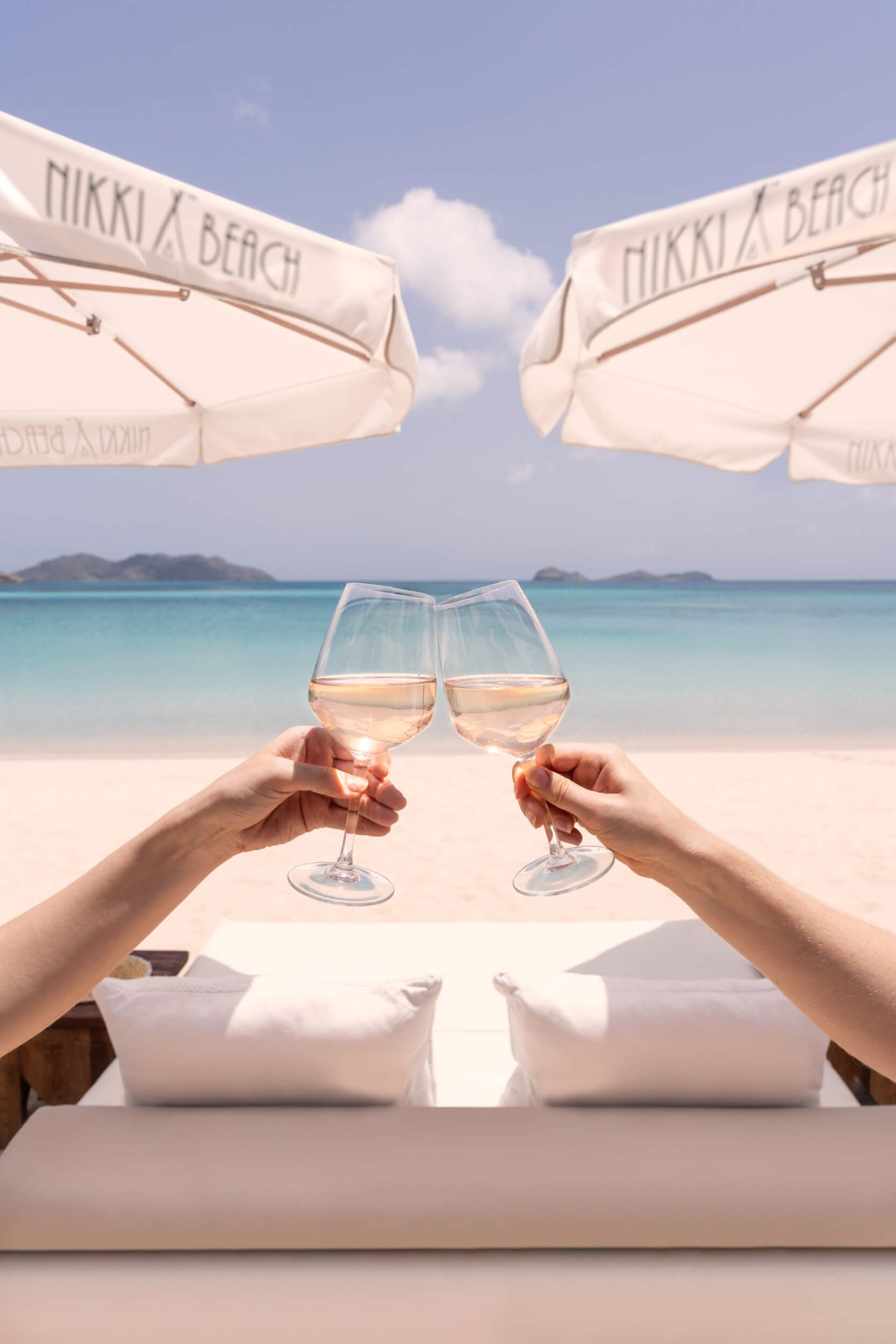 Two women toasting two glasses on the beach with the sea and parasols in the background