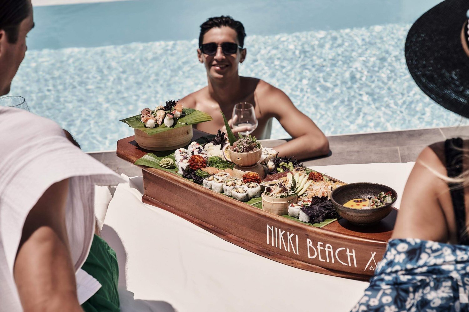 People around a pool enjoying a sushi platter, served on a wooden boat shaped board