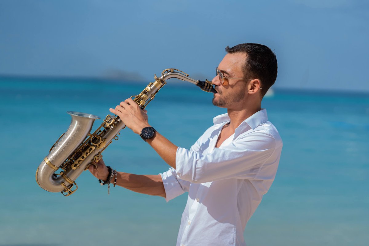 Man in a white shirt playing the saxaphone with the sea in the background