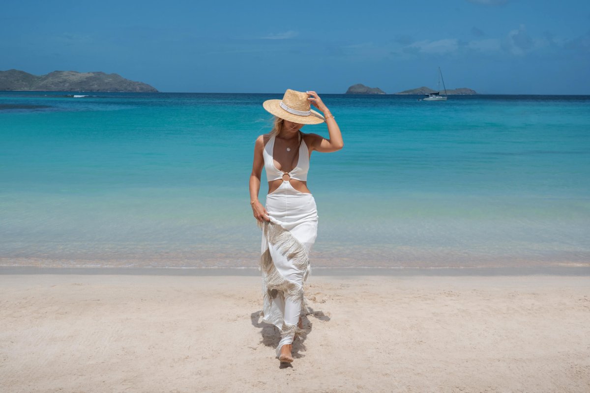 Women in white dress holding her straw hat on her head with blue sea behind her