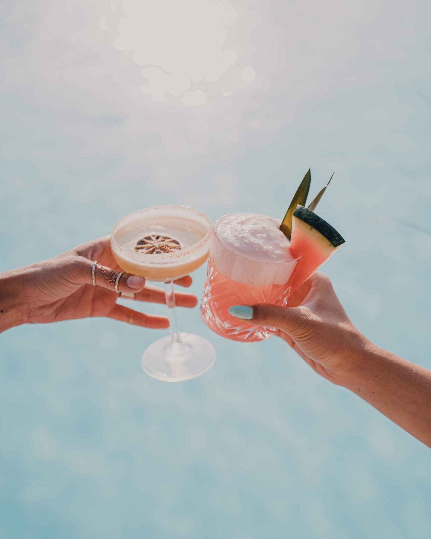 Two hands cheersing tropical cocktails garnished with watermelon and orange