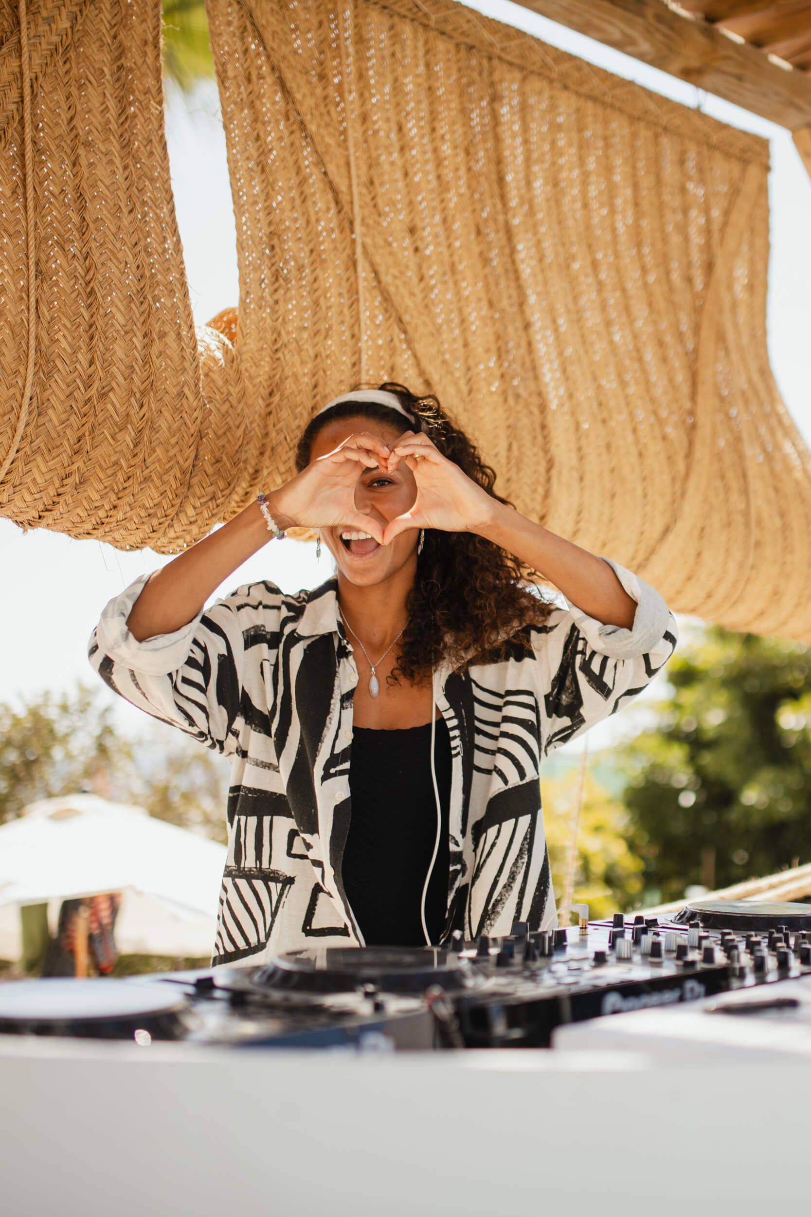 A women DJ in front of her mixing decks, making a heart shape with her hands at the camera