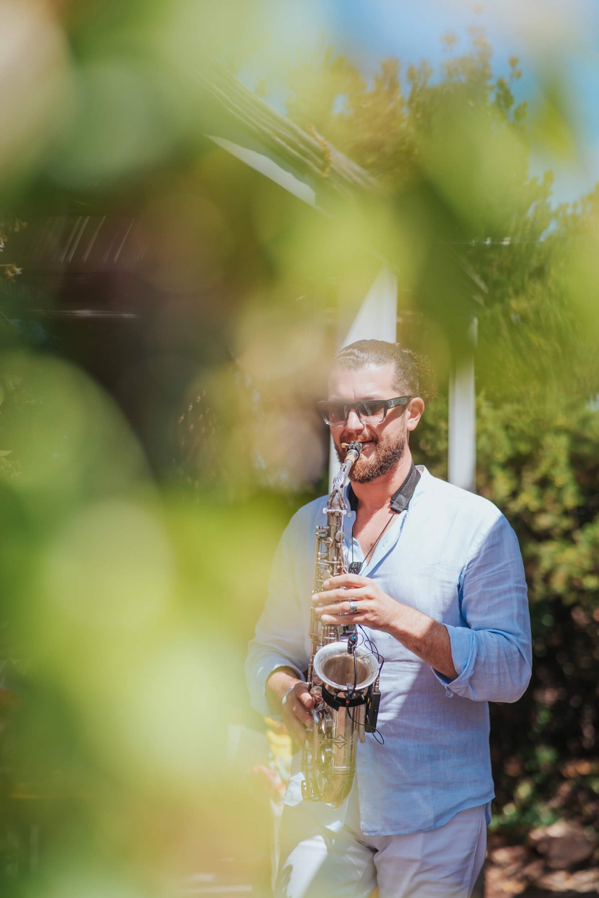 Man in a light blue shirt playing the saxophone with leaves in the foreground