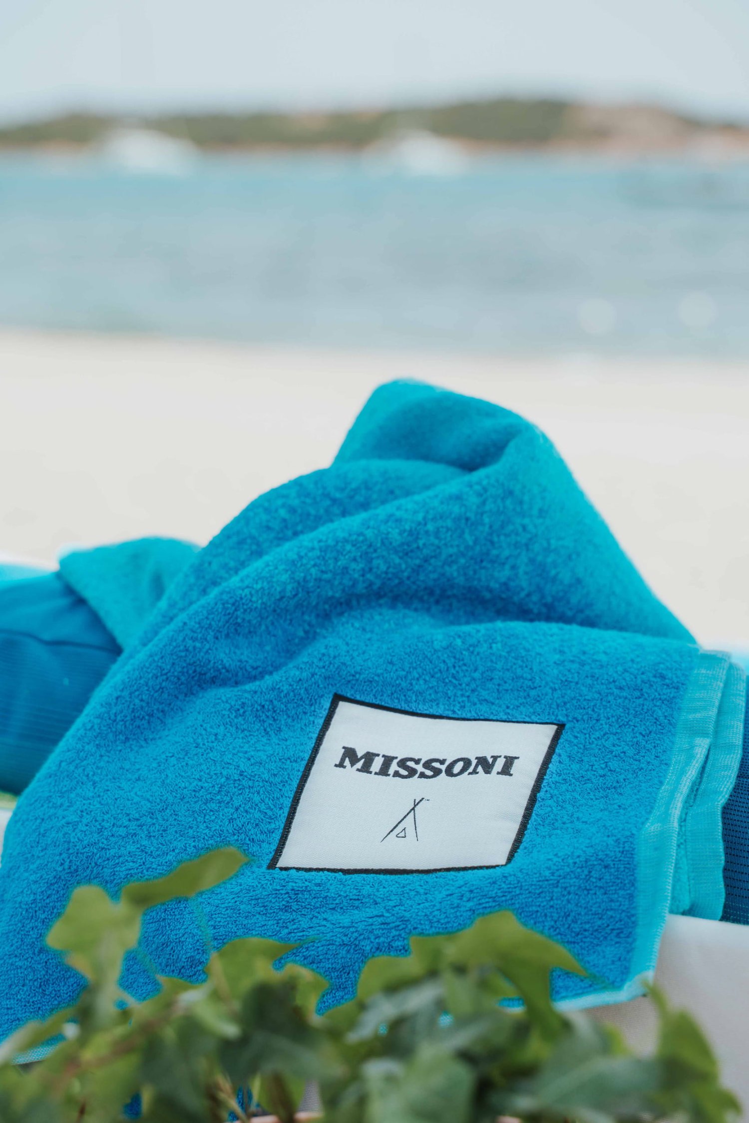 A close-up view of a vibrant turquoise Missoni and Nikki Beach co-branded towel resting on a sunbed. Framed by green foliage in the foreground and a blurred sandy beach and ocean in the background, this image highlights the exclusive lifestyle fashion collaborations and experiential marketing activations hosted at Nikki Beach Resort & Spa Koh Samui