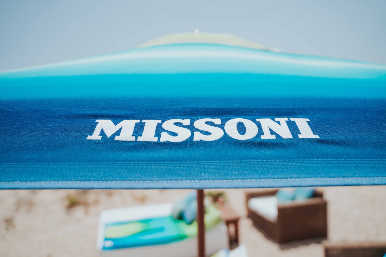 A close-up view of a vibrant blue and turquoise gradient beach umbrella prominently displaying the white Missoni logo. With a blurred sandy beach and matching colorful lounge beds in the background, this image perfectly illustrates the impactful, high-visibility pop-ups and luxury brand collaborations hosted at Nikki Beach Resort & Spa Koh Samui
