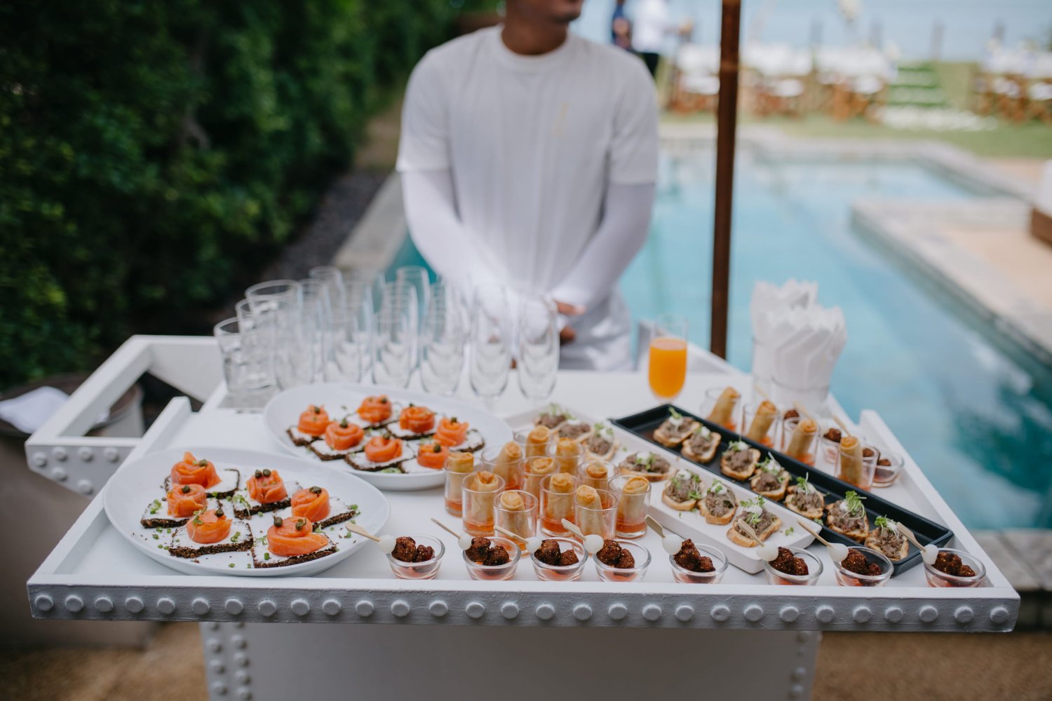 A close-up view of an elegant white catering cart filled with beautifully presented appetizers, including smoked salmon canapés, spring rolls in glass cups, and bite-sized hors d'oeuvres, alongside rows of champagne flutes. A server in a white uniform stands ready in the blurred background next to a tranquil resort pool, highlighting the seamless, high-quality end-to-end event delivery and dining experiences offered for corporate events at Nikki Beach Resort & Spa Koh Samui