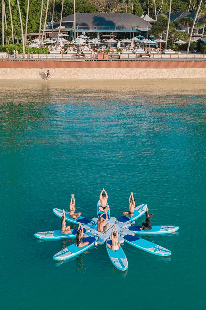 An aerial view of a team-building wellness session on the bright turquoise ocean. A group of people sits on blue and white paddleboards arranged in a star formation, practicing a seated yoga pose with their arms raised together. The sandy beach and vibrant Nikki Beach Resort & Spa Koh Samui, complete with palm trees and signature white parasols, serve as a stunning backdrop, perfectly illustrating the seamless integration of business retreats and lifestyle-driven island experiences