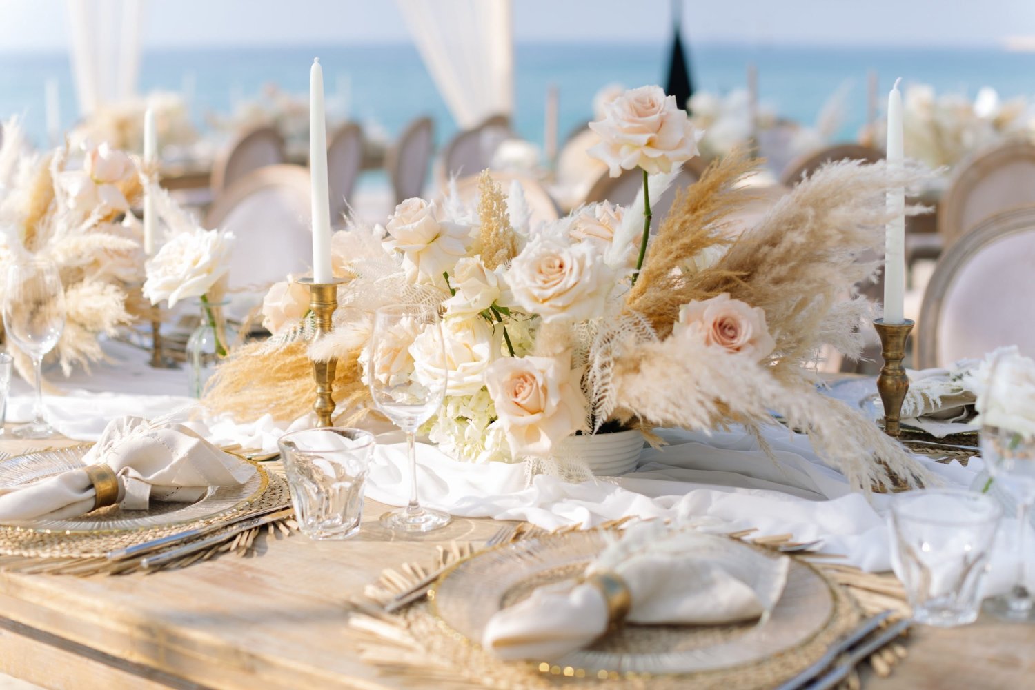A close-up of a beautifully styled outdoor wedding table at Nikki Beach Resort & Spa Koh Samui. The elegant wooden table features a romantic, bohemian centerpiece of pale blush roses and fluffy pampas grass, surrounded by tall white taper candles in brass holders. Woven placemats, gold-rimmed glass chargers, and folded linen napkins complete the luxurious coastal setting, with the blue ocean gently blurred in the background