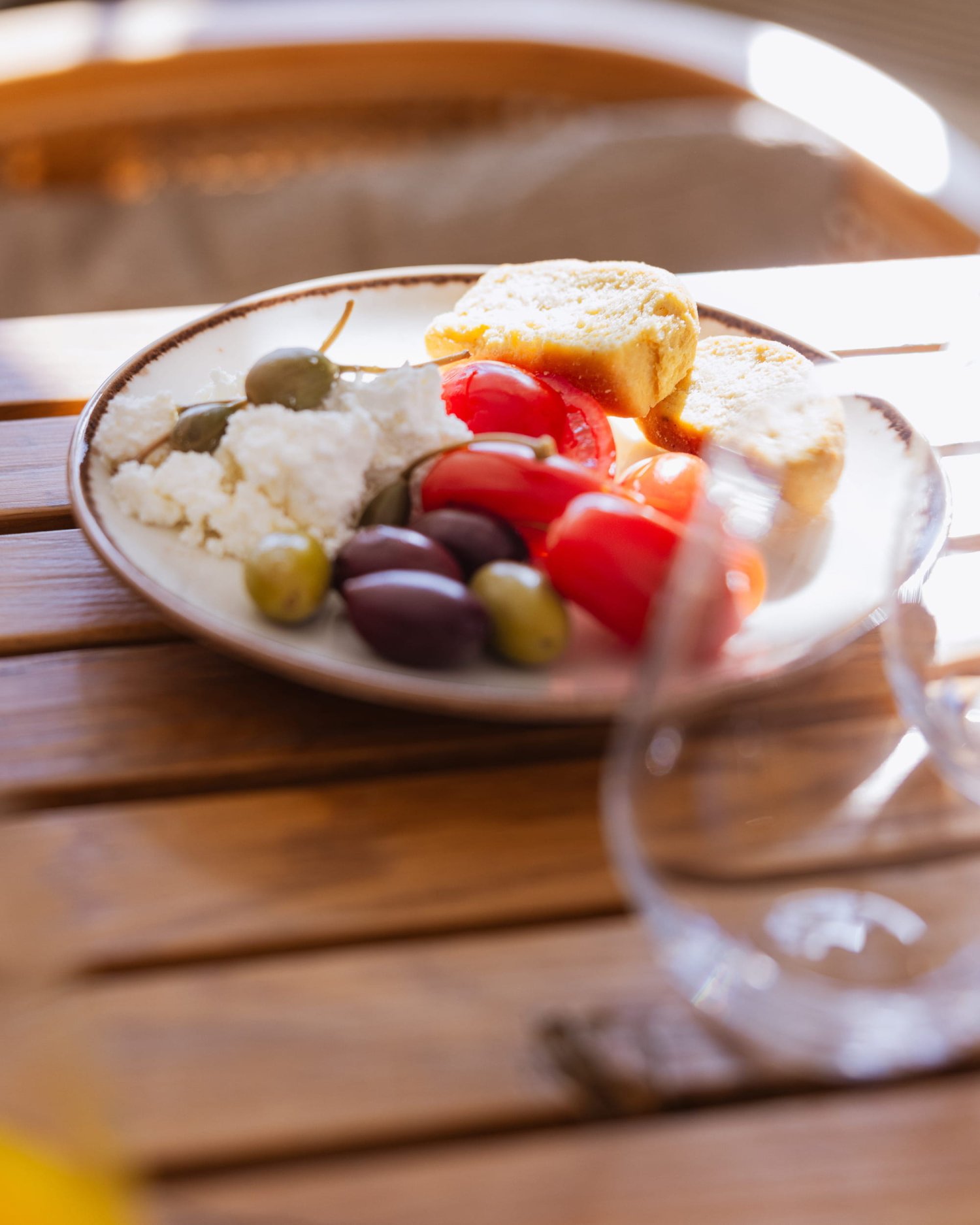 A sunlit, close-up view of a fresh Mediterranean breakfast plate featuring crumbled feta cheese, plump Kalamata olives, bright red tomatoes, capers, and crusty bread, resting on a wooden table to highlight the locally sourced ingredients at Nikki Beach Santorini