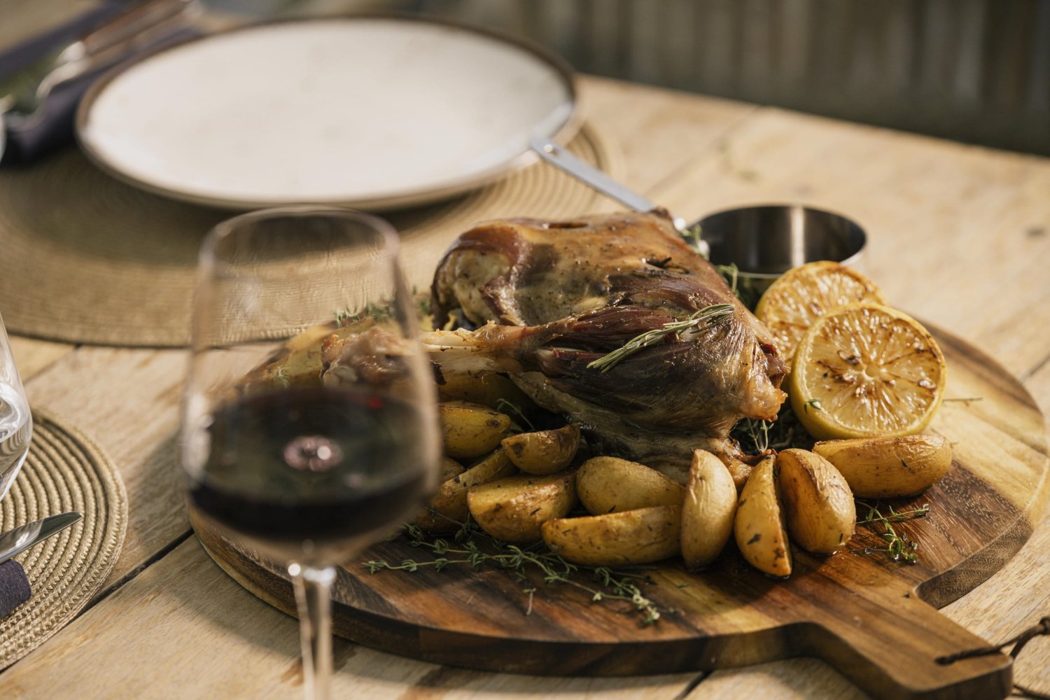 A rustic wooden serving board presenting a beautifully roasted leg of lamb surrounded by seasoned potato wedges, fresh herbs, and charred lemon halves. Accompanied by a blurred glass of red wine, this hearty dish showcases the extraordinary culinary innovation crafted by the executive chefs at Nikki Beach Santorini