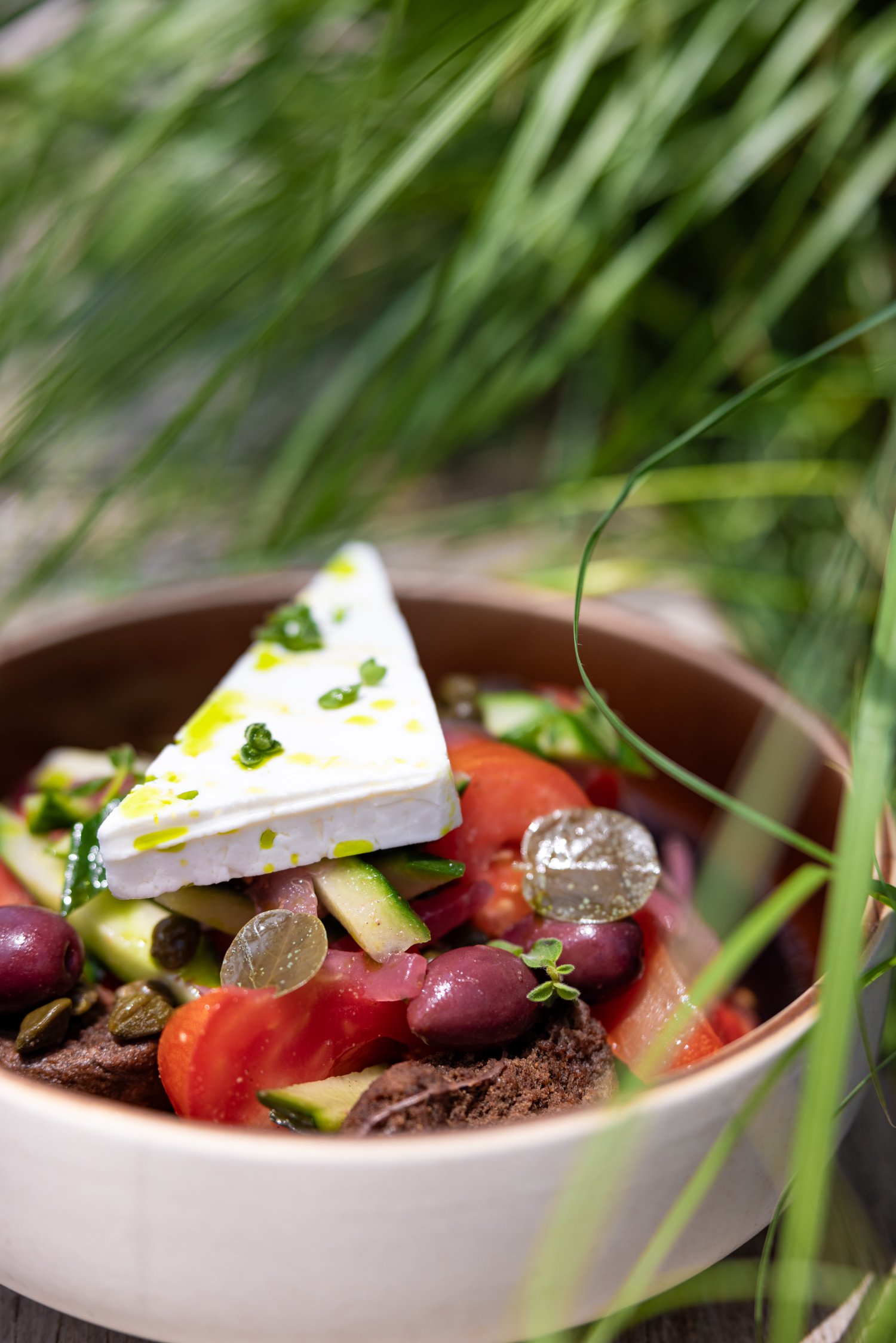 A close-up view of a vibrant Greek salad served in a bowl, featuring a thick triangular slice of feta cheese drizzled with olive oil and herbs, resting atop fresh tomatoes, cucumbers, olives, capers, and traditional rusks. Framed by blurred green foliage, this dish perfectly highlights the fresh, local ingredients celebrated in the resort's menu