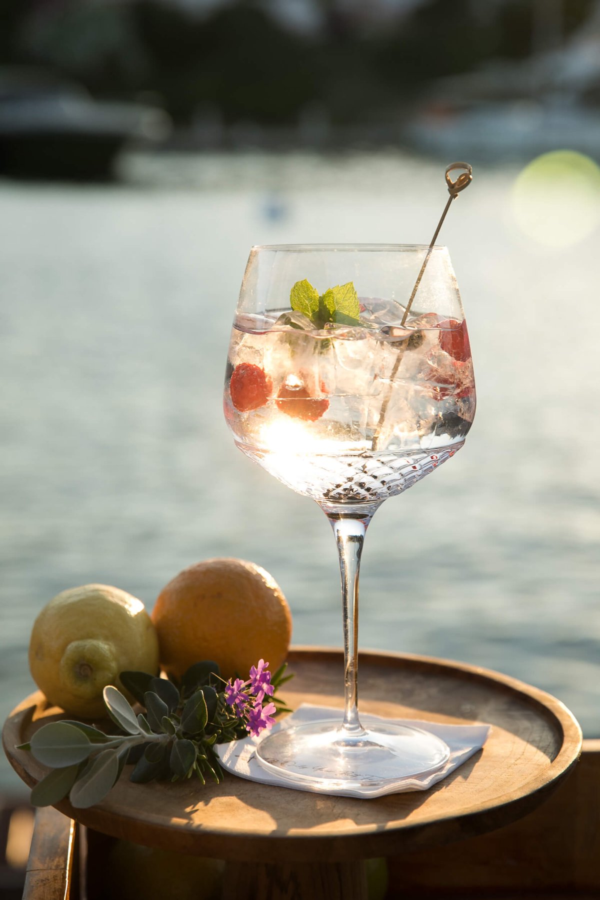Cocktail in a large glass on a wooden stool with fruit and garnish next to it and the sea in the background