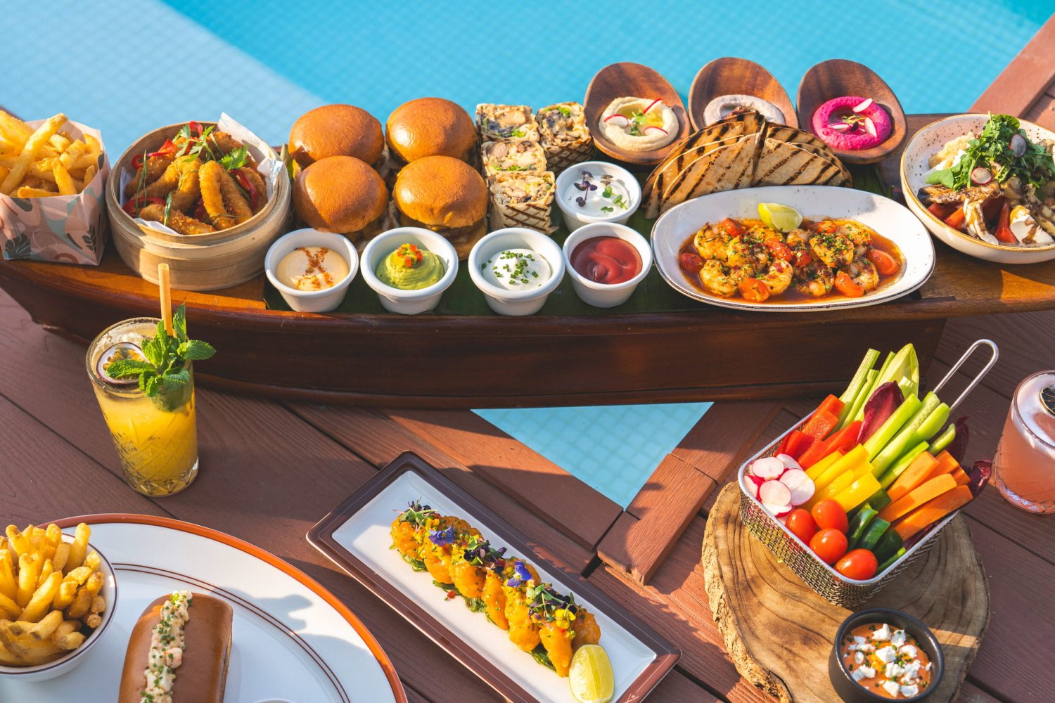 A close-up view of the signature Culinary Cravings sharing boat, a large wooden boat-shaped platter filled with an appetizing assortment of brunch items including sliders, wraps, calamari, shrimp, and various dips. The feast is accompanied by fresh vegetable sticks, fries, and vibrant cocktails, set on a wooden deck directly next to a bright blue swimming pool, perfectly capturing the indulgent Saturday poolside brunch experience