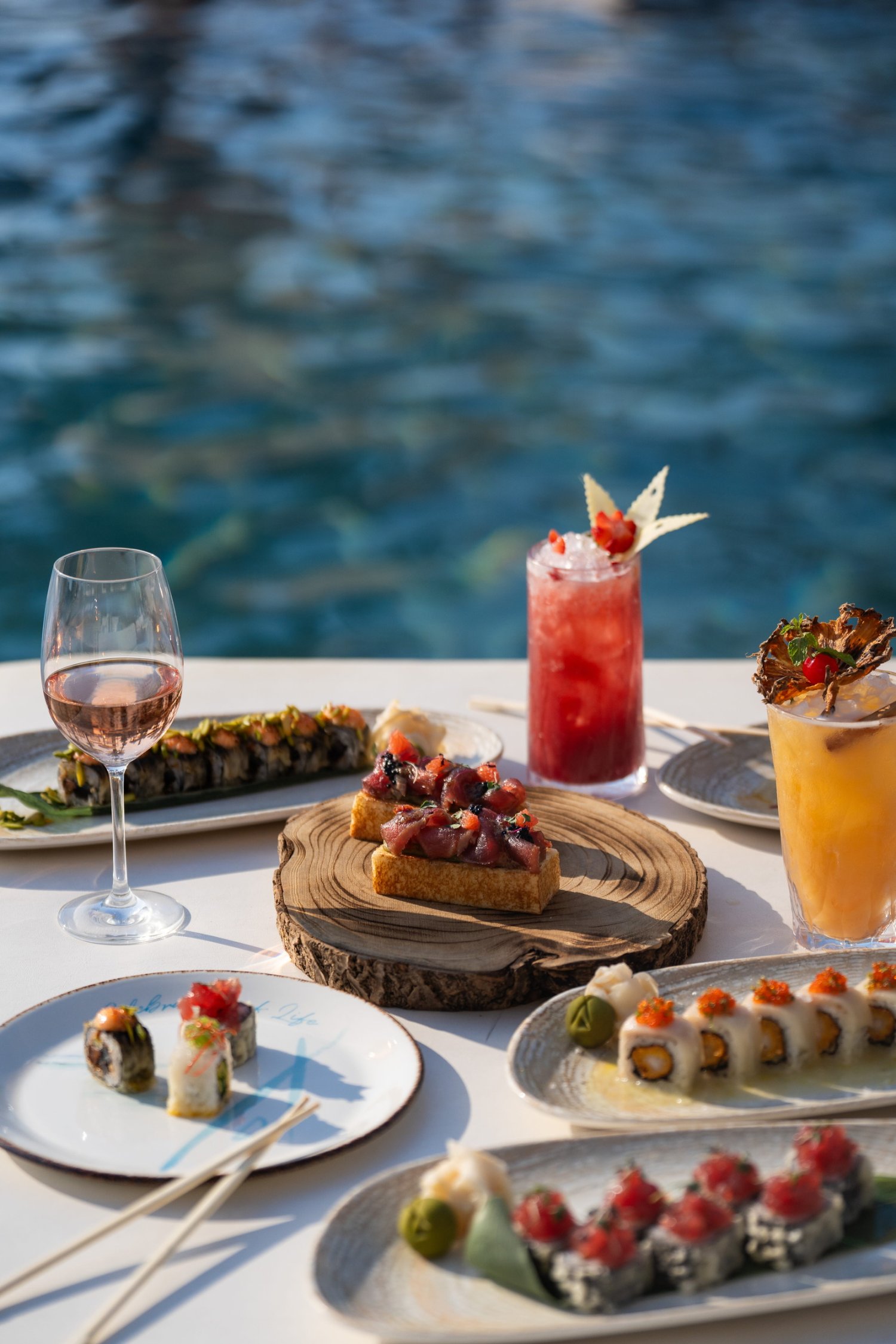 A close-up view of an inviting brunch spread set on a white table directly beside sparkling blue pool water. The feast features an array of fresh sushi rolls and tuna tartare served on thick toast atop a rustic wooden board, perfectly paired with a glass of rosé and two vibrant, expertly crafted cocktails. This mouth-watering scene beautifully captures the reimagined menu, fresh seafood, and vibrant daytime atmosphere of the iconic Amazing Sundays Brunch